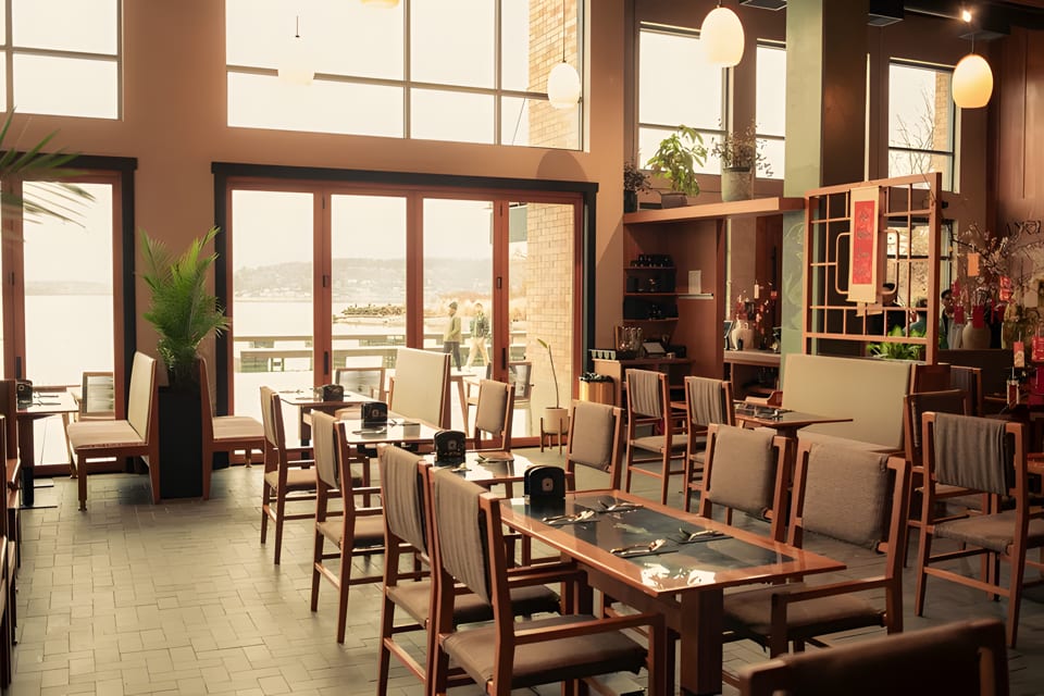 A restaurant dining room with wooden tables and chairs, large windows overlooking a body of water.