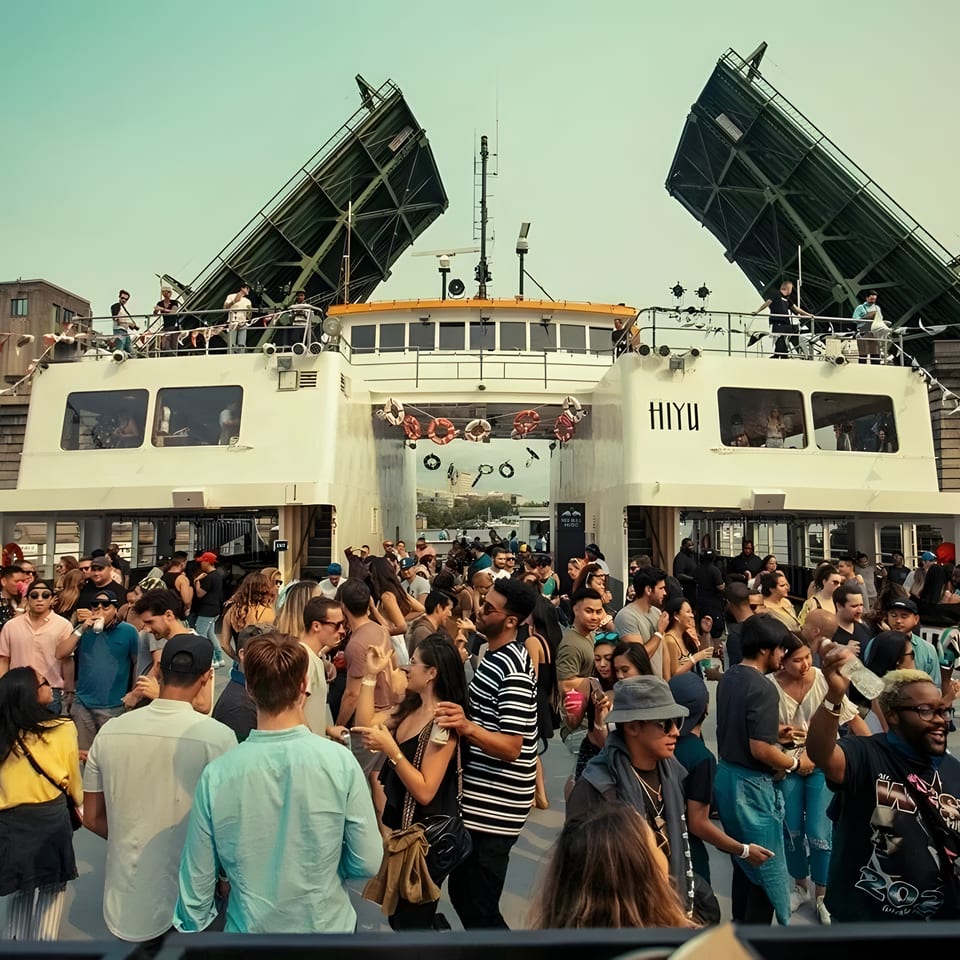 A lively crowd of people on a boat party, with a bridge in the background.
