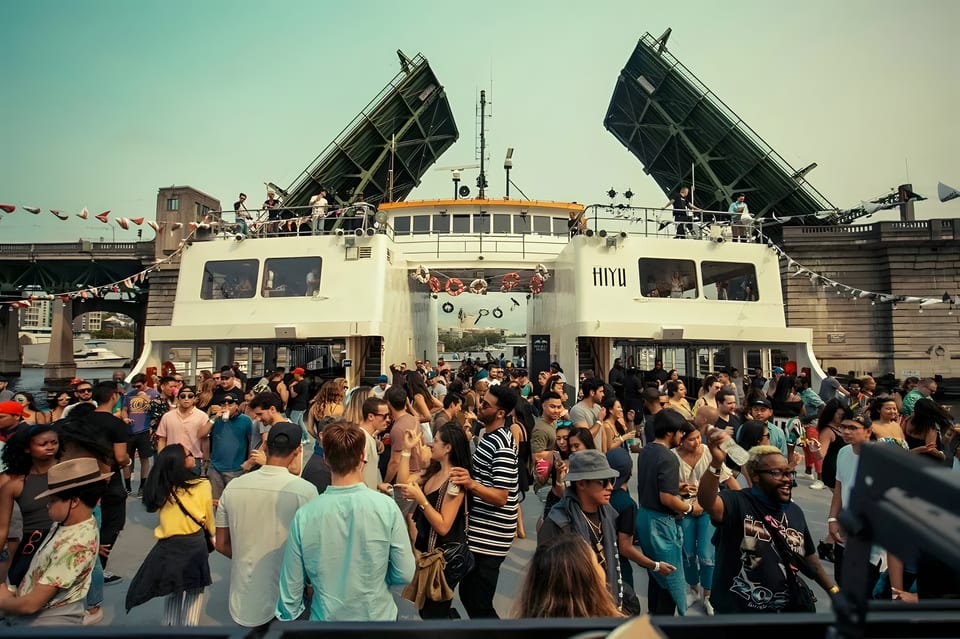 A lively crowd of people on a boat party, with a bridge in the background.