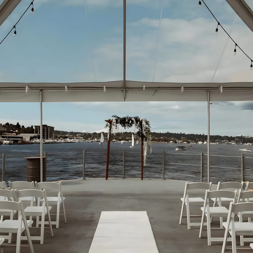 Outdoor wedding ceremony setup with white chairs, floral arch, and waterfront view under a clear tent.