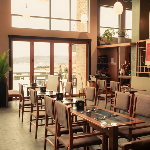 Spacious restaurant dining room with wooden tables, chairs, and large windows overlooking a body of water.