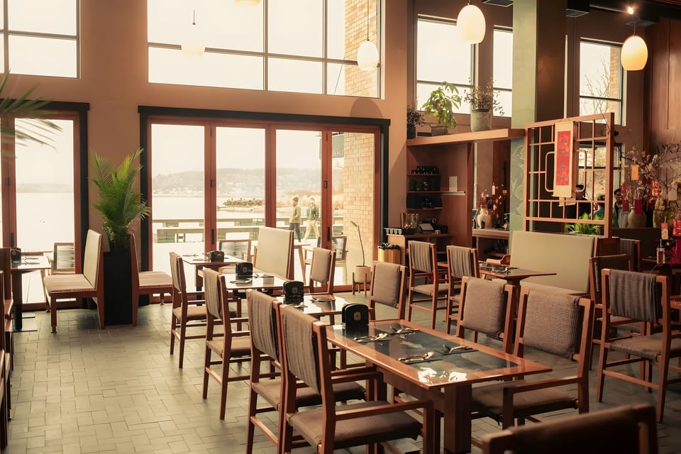 Spacious restaurant dining room with wooden tables, chairs, and large windows overlooking a body of water.