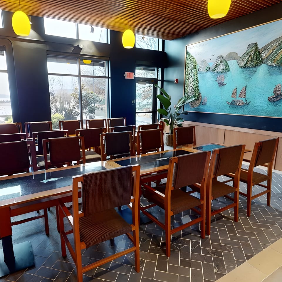 A private dining room with a long table, chairs, large windows, and a vibrant seascape painting.