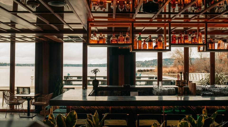 A modern bar with a wooden ceiling, liquor bottles, and large windows overlooking a lake.