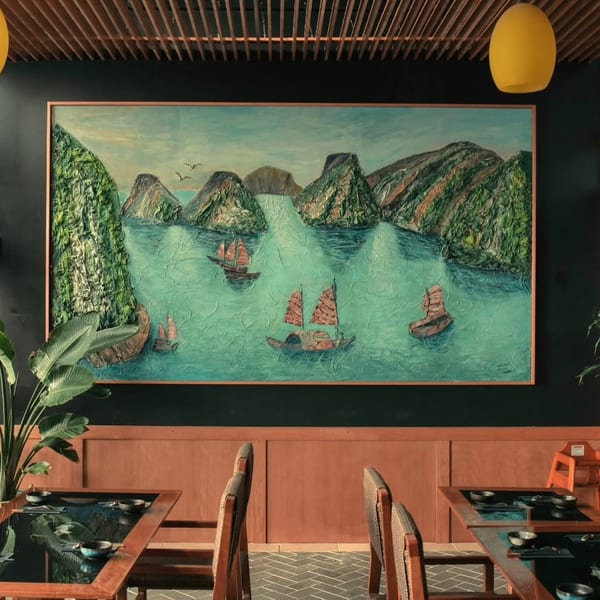 Restaurant interior with a large textured painting of boats on water, wooden tables, and plants.