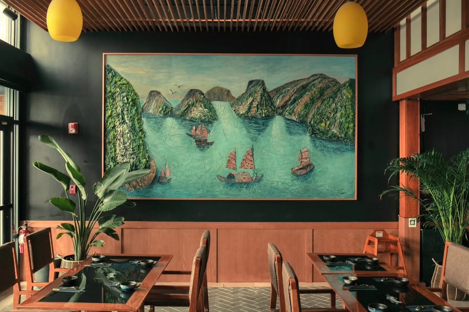 Restaurant interior with a large textured painting of boats on water, wooden tables, and plants.