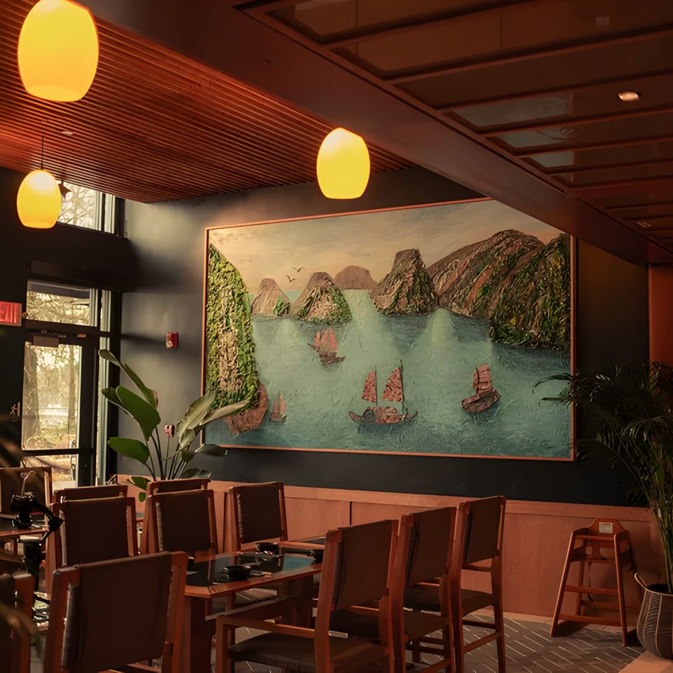 Restaurant dining room with a large painting of boats on water, wooden tables, and warm lighting.