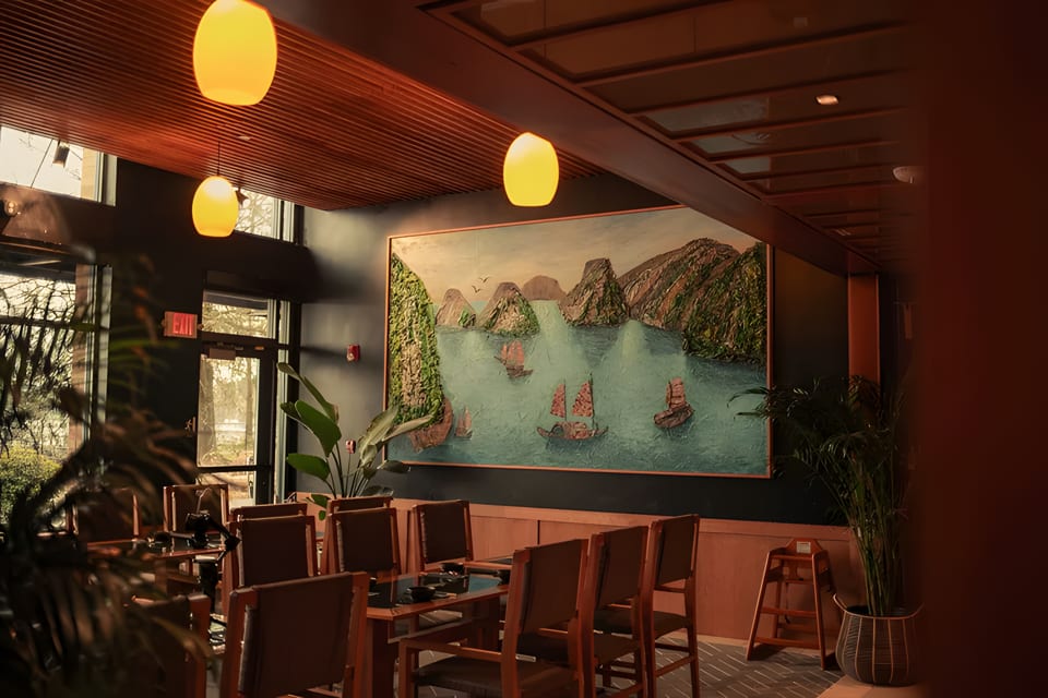 Restaurant dining room with a large painting of boats on water, wooden tables, and warm lighting.