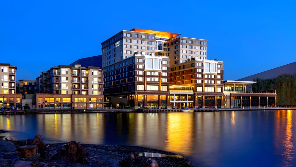 A large, illuminated Hyatt Regency hotel and other buildings line a waterfront at dusk.