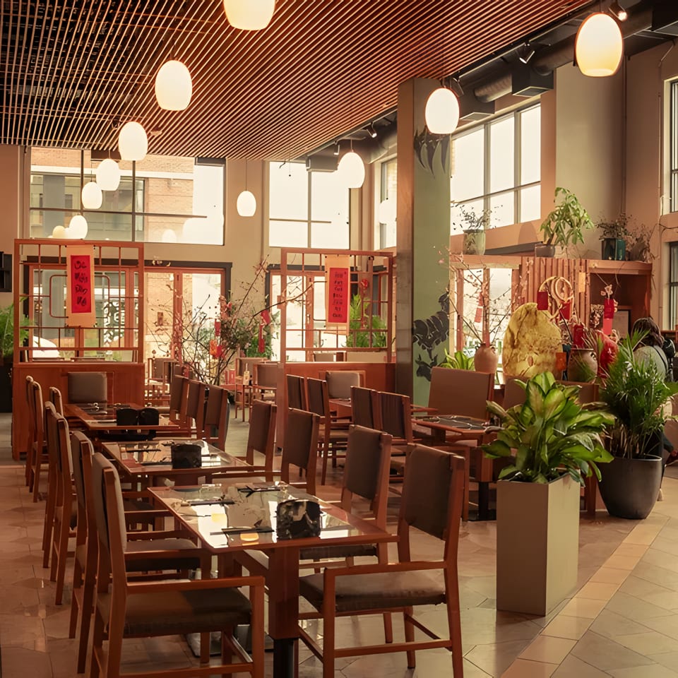 Spacious restaurant dining room with wooden tables and chairs, large windows, and decorative plants.