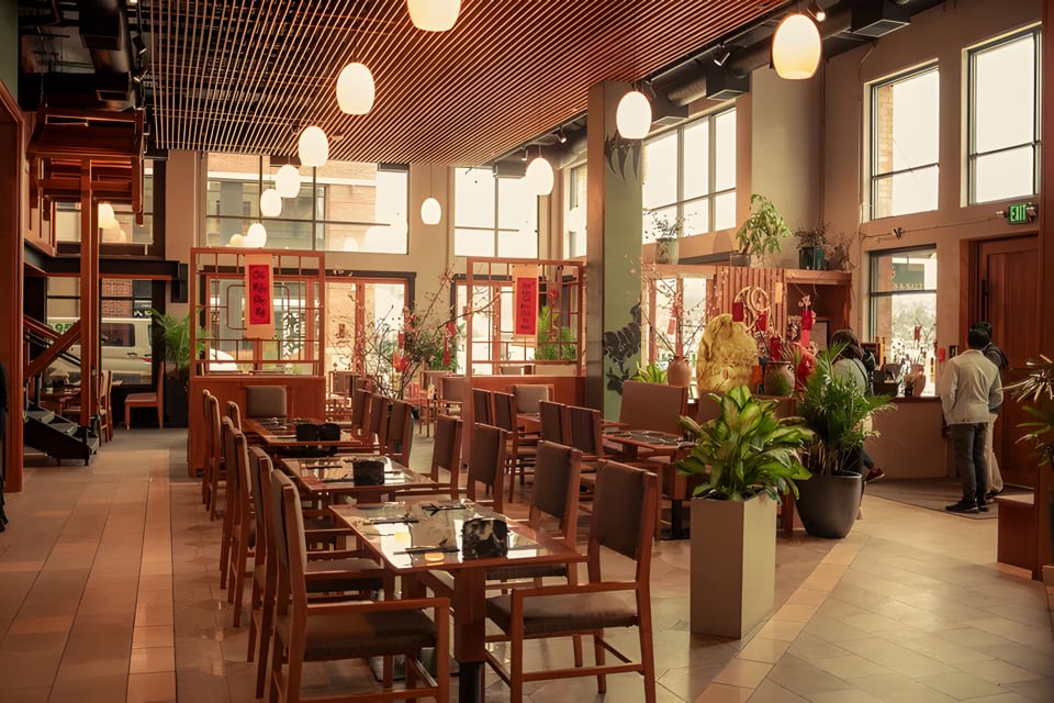 Spacious restaurant dining room with wooden tables and chairs, large windows, and decorative plants.