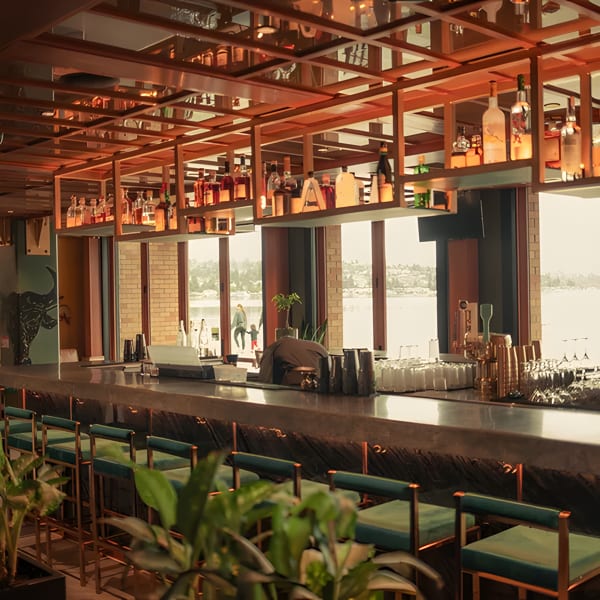 A modern bar with a long counter, teal stools, and bottles on shelves, overlooking a body of water.