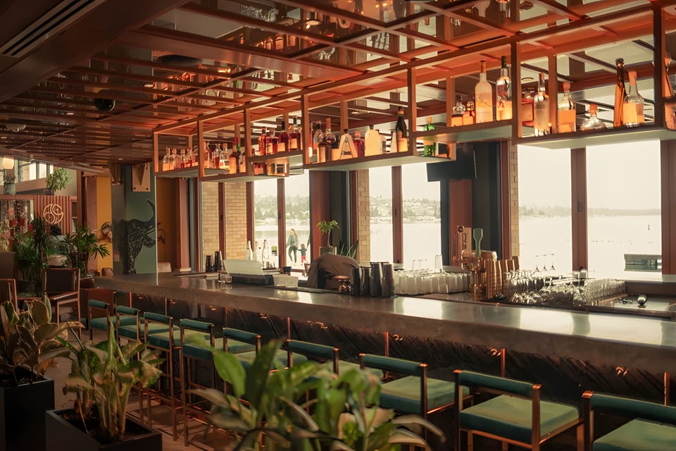 A modern bar with a long counter, teal stools, and bottles on shelves, overlooking a body of water.